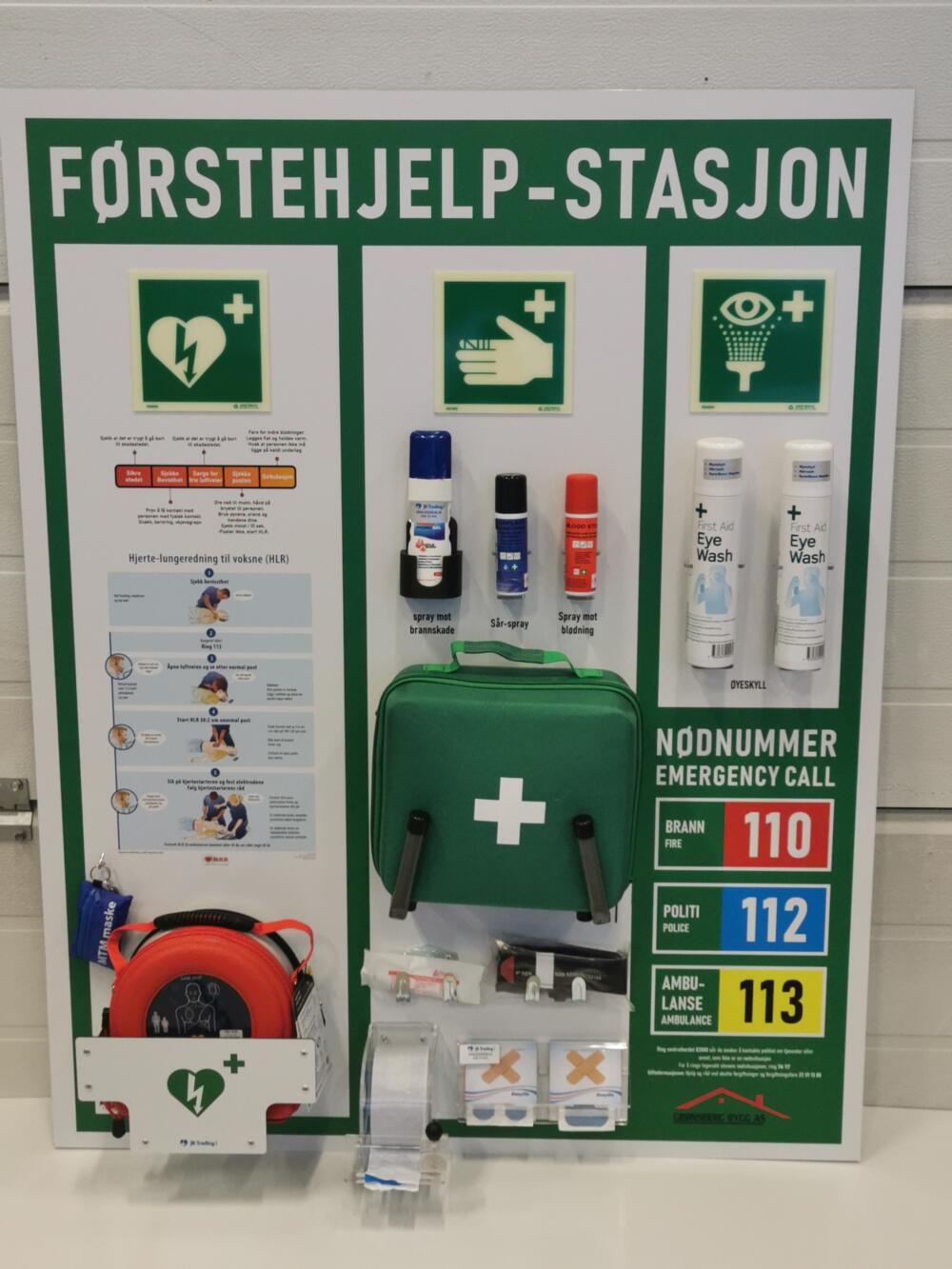Grønn HMS-tavle merket Førstehjelp-stasjon, med beredskapsveske, sprayer, øyeskyll, hjertestarter og nødnummer 110, 112 og 113